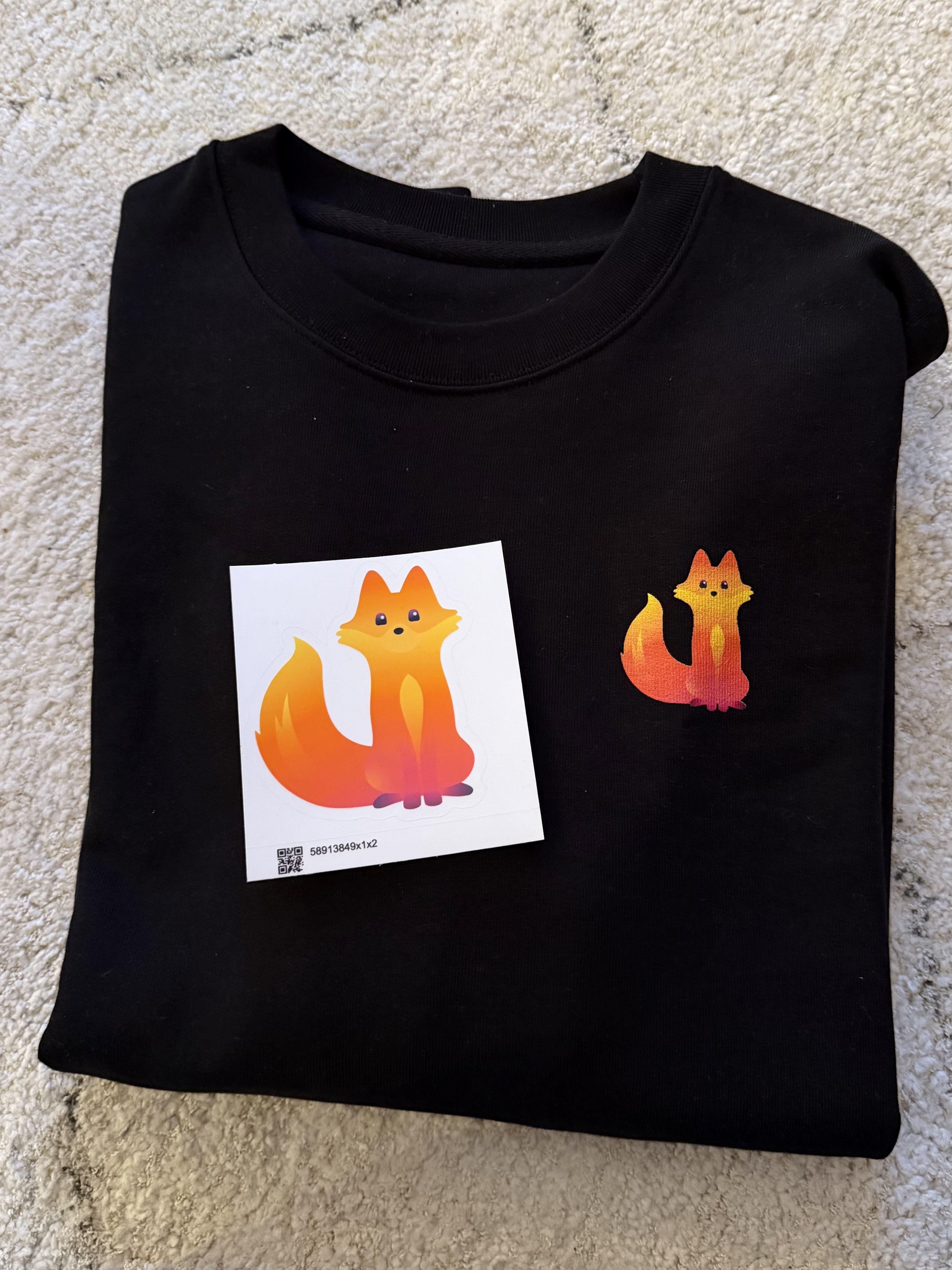Image showing some of the the new Firefox merch, including a vinyl sticker and a black shirt. Both feature the new Mascot "Kit"