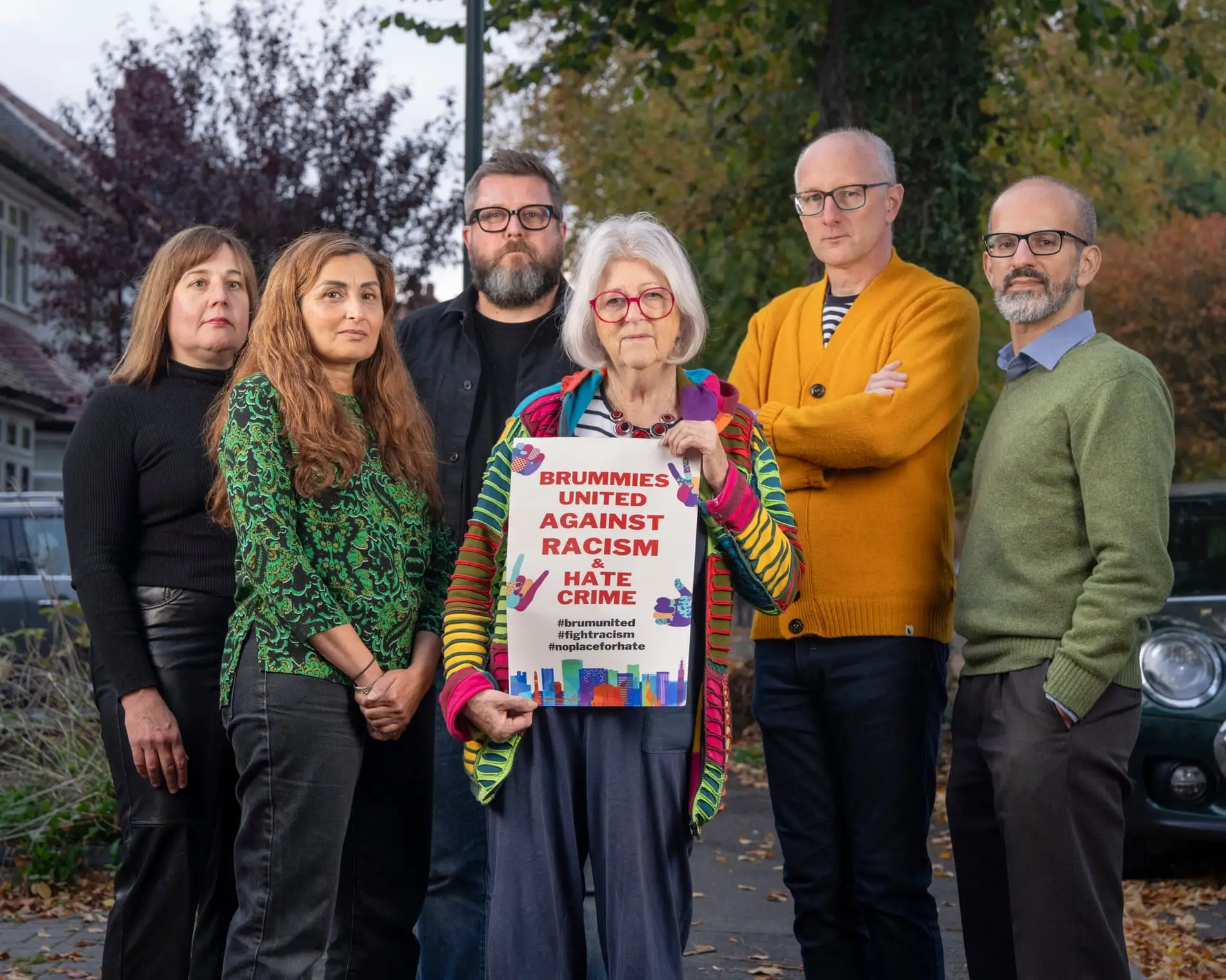 Six neighbours (three men and three women) standing on a pavement in their neighbourhood. The woman in the middle of the photo is holding a sign which says "Brummies united against racism & hate crime", with three hashtags underneath: "#brumunited", "#fightracism", and "#noplaceforhate".