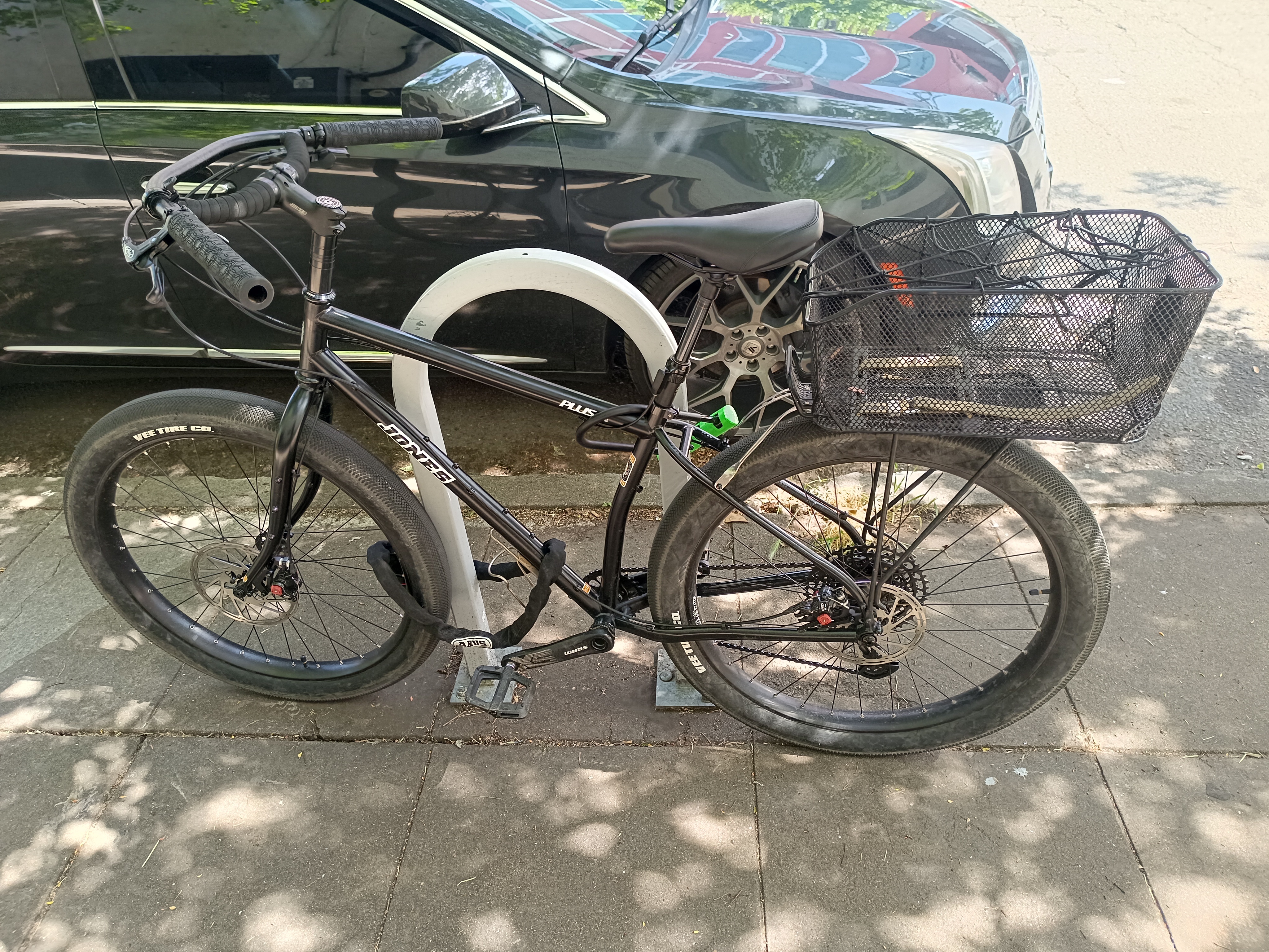 Black bicycle leaning on bicycle rack with a metal mesh basket on back, in front of a car