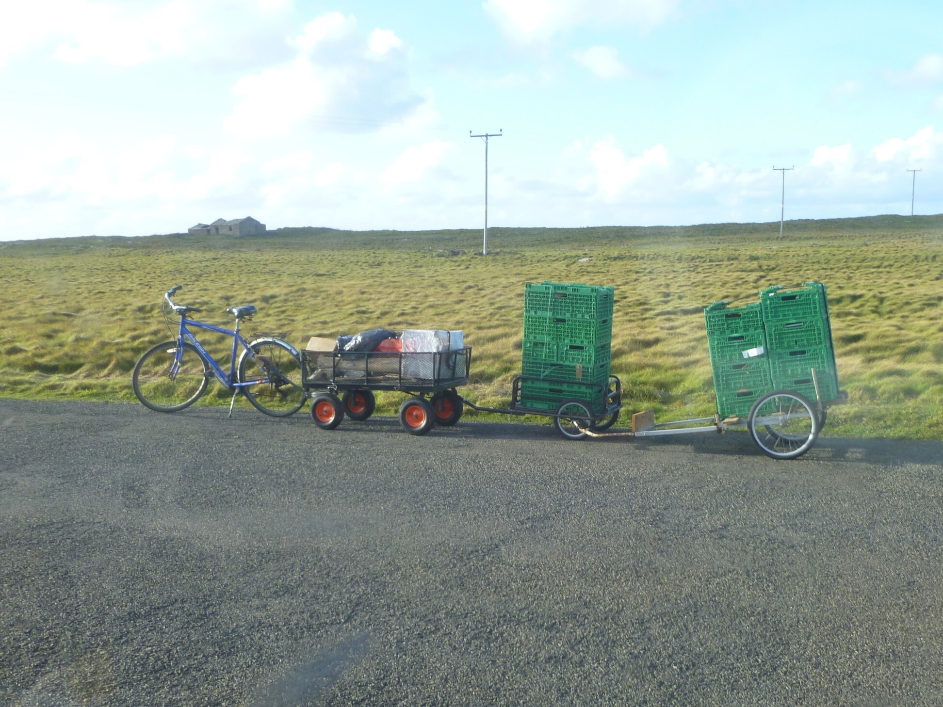 The bike train on a road, carrying rather tall stacks of green crates, as well as other miscellaneous items