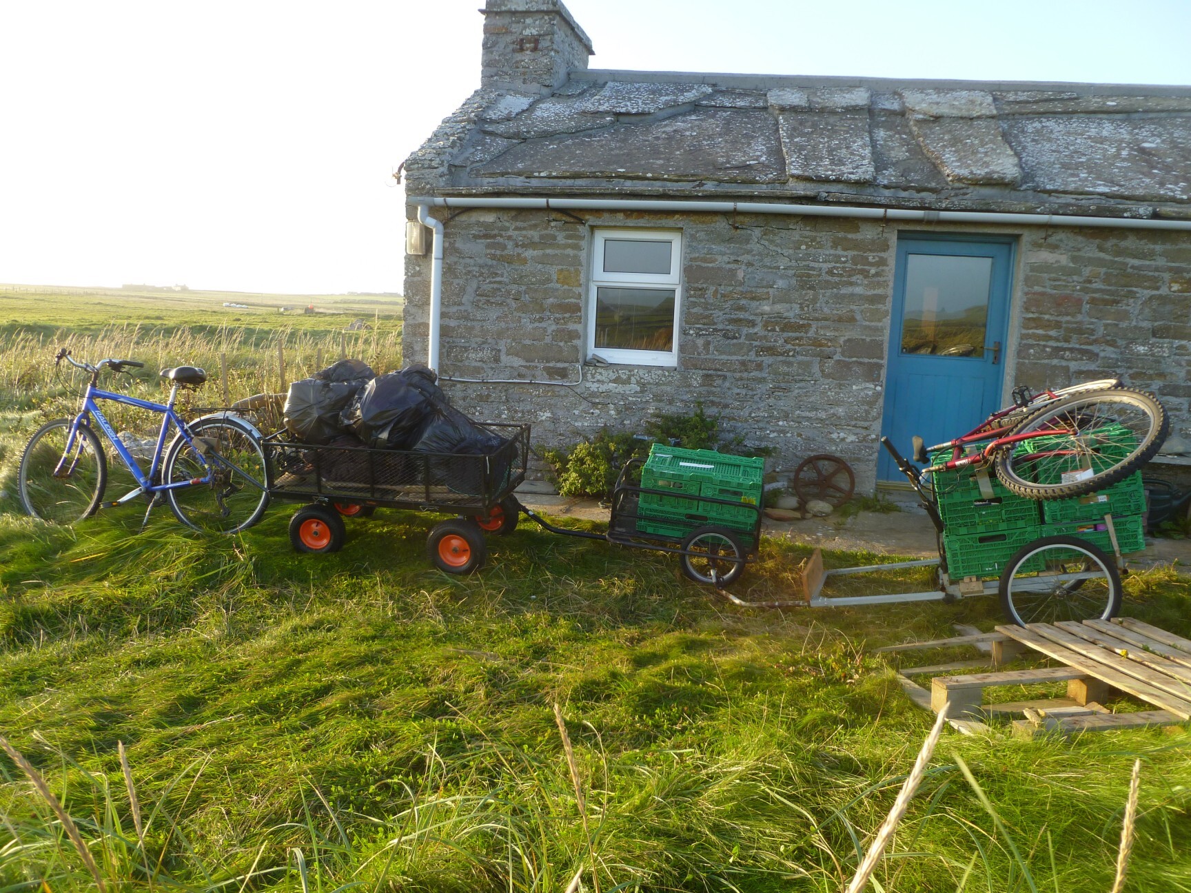 In front of a stone house with a blue door. The trailers hold bin bags and green crates with another bicycle laid on top
