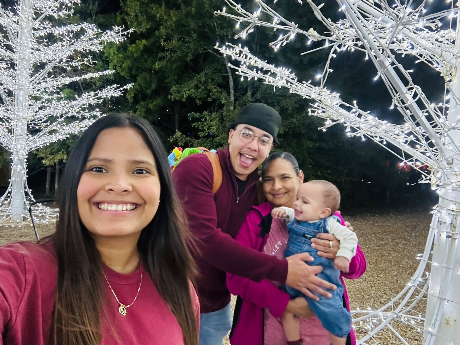 A woman standing amid white metal trees with Christmas lights takes a photo while a man  and older woman holding a baby pose behind her